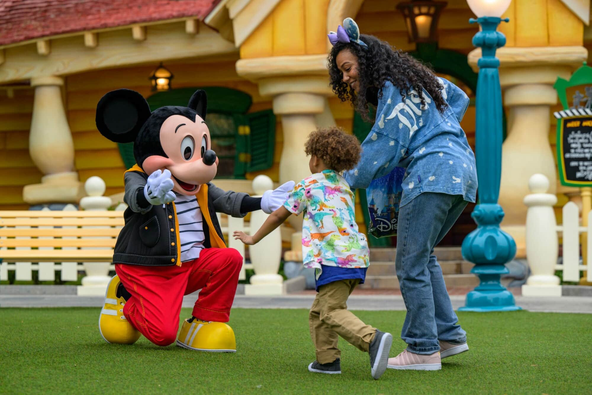 A woman and small child excitedly greet Mickey Mouse in front of his house, with Mickey's arms outstretched to say hi.