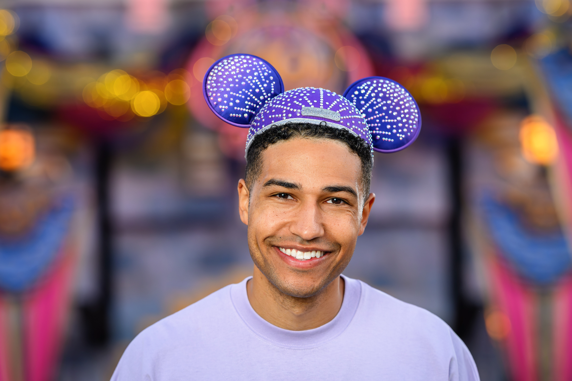 2026DLRFinaleMerchCollection1 | The Main Street Mouse Young Man Wearing Purple Dotted Minnie Mouse Ears and Smiling at a Festive Outdoor Event with Colorful Bokeh Lights Behind Him