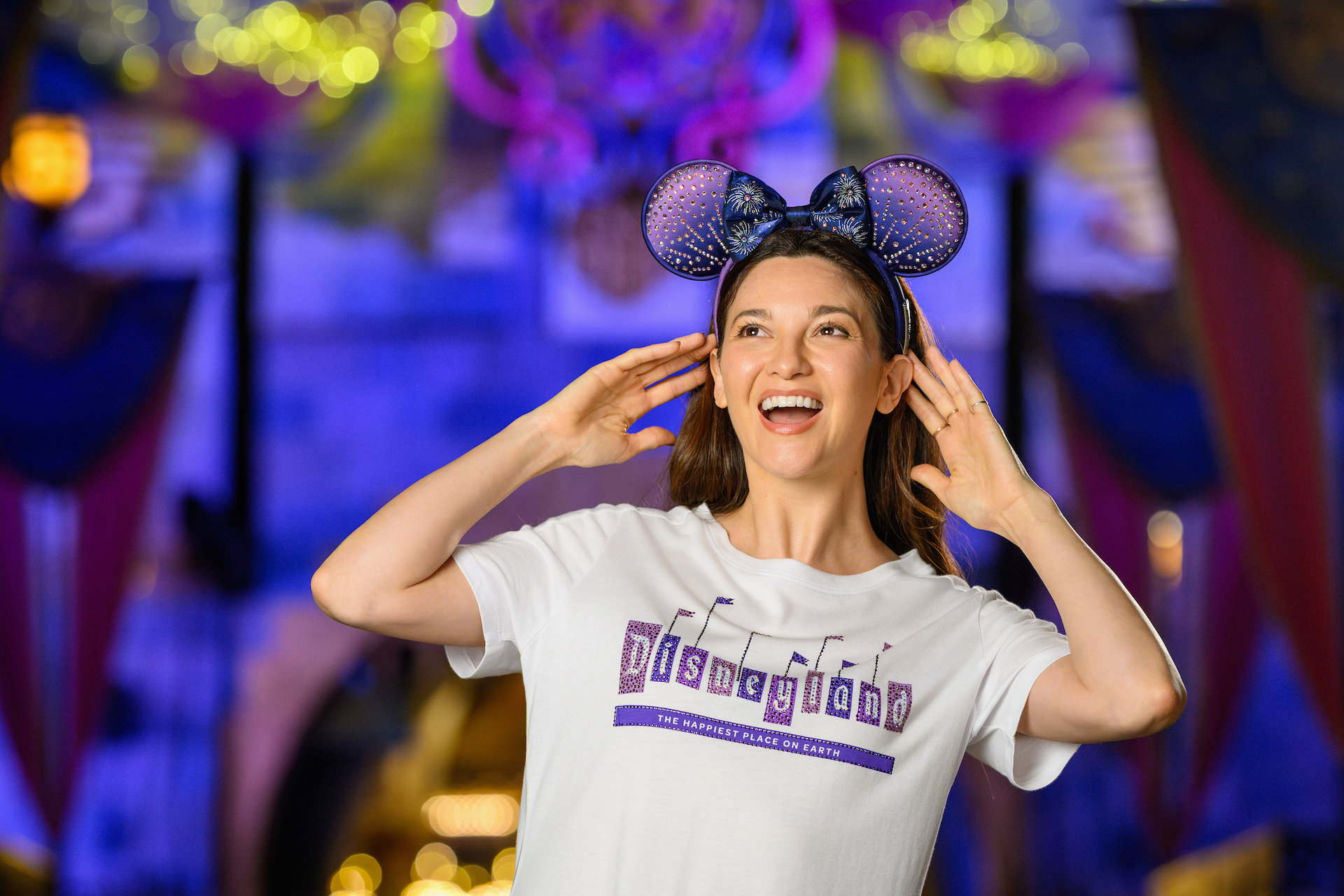 2026DLRFinaleMerchCollection2 | The Main Street Mouse Smiling Woman Wearing Purple Glitter Minnie style Ears Headband Hands Near Her Ears Colorful Lights in the Background