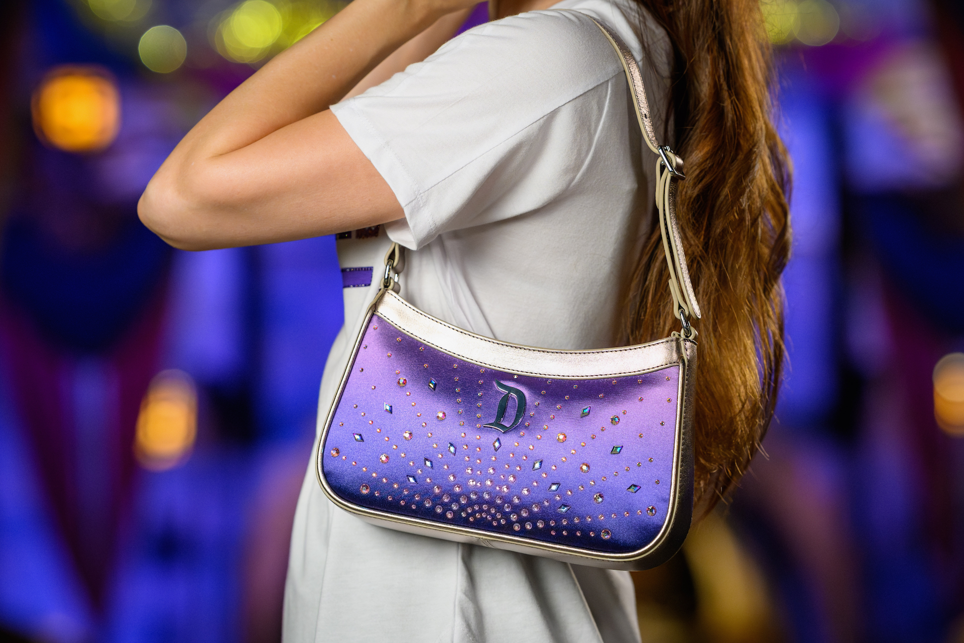 2026DLRFinaleMerchCollection3 | The Main Street Mouse Person in a Light Gray Shirt Carries a Purple Rhinestone embellished Purse over the Shoulder Against a Colorful Blurred Background