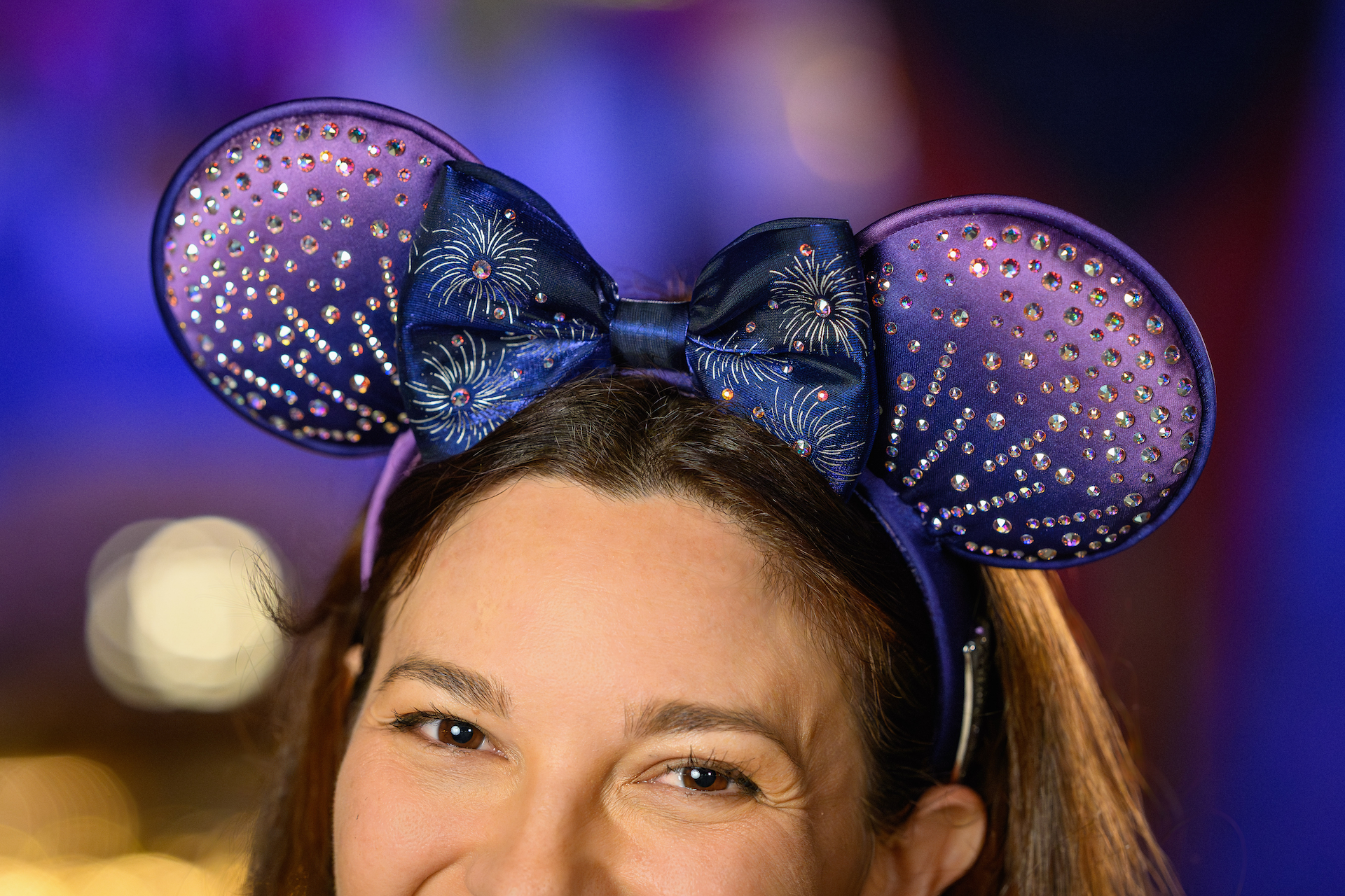 2026DLRFinaleMerchCollection4 | The Main Street Mouse Woman Wearing a Purple Rhinestone studded Minnie Mouse Ears Headband with a Blue Bow Smiling at a Celebration