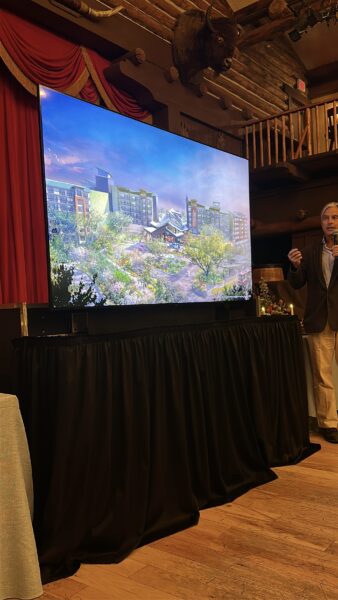 IMG1306 | The Main Street Mouse Presenter speaks beside a large screen displaying a modern architectural rendering in a rustic lodge like venue with red curtains and a mounted elk head above