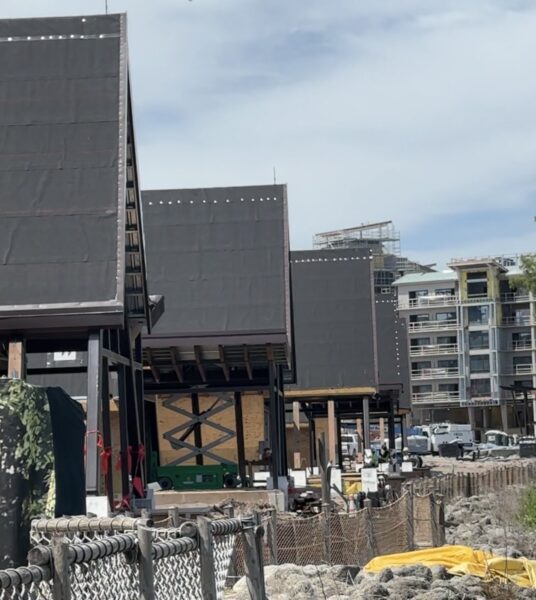 Screenshot | The Main Street Mouse Row of dark modular building units on steel supports under construction along a body of water with a chain link fence and construction debris in the foreground and residential buildings in the background