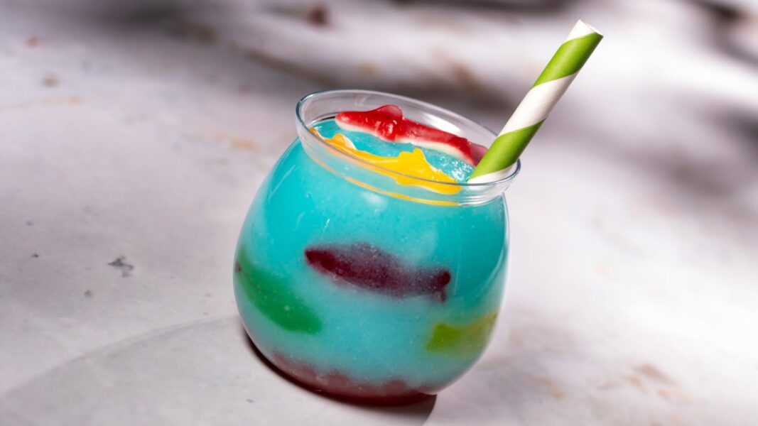IMG1711 | The Main Street Mouse Blue Tropical Drink in a Curved Glass with a Green and white Striped Straw and Red yellow Garnish on a Light Surface