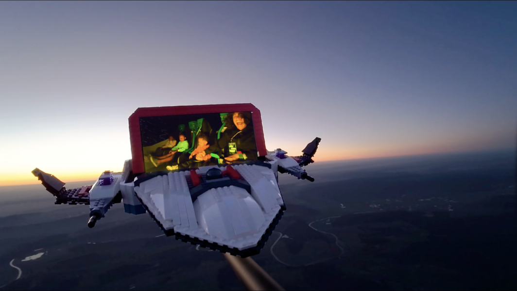 LEGO spaceship with a redframed screen showing smiling people flying over a coastline at sunset | The Main Street Mouse LEGO spaceship with a red framed screen showing smiling people flying over a coastline at sunset