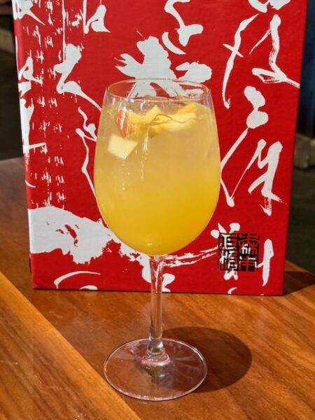 Sake Sangria 2 | The Main Street Mouse Orange Cocktail with Ice in a Stemmed Glass Lemon Twist Garnish on a Wooden Table with a Red Abstract Backdrop