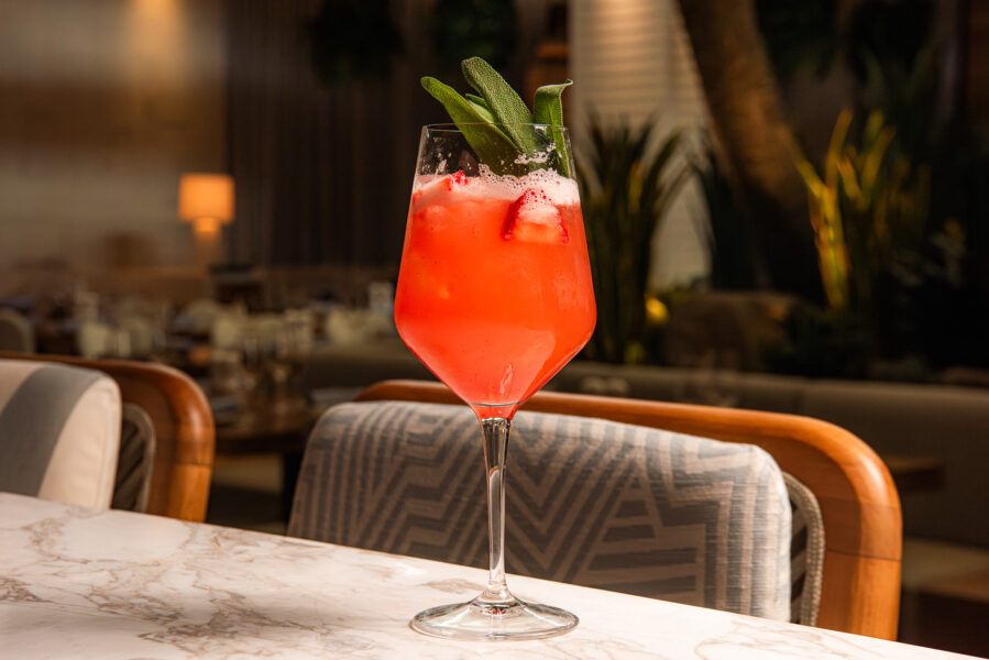 Strawberry FieldsNA CocktailSHOL | The Main Street Mouse Pink Cocktail in a Stemmed Glass with Ice and a Green Herb Garnish on a Marble Table Restaurant Setting in the Background