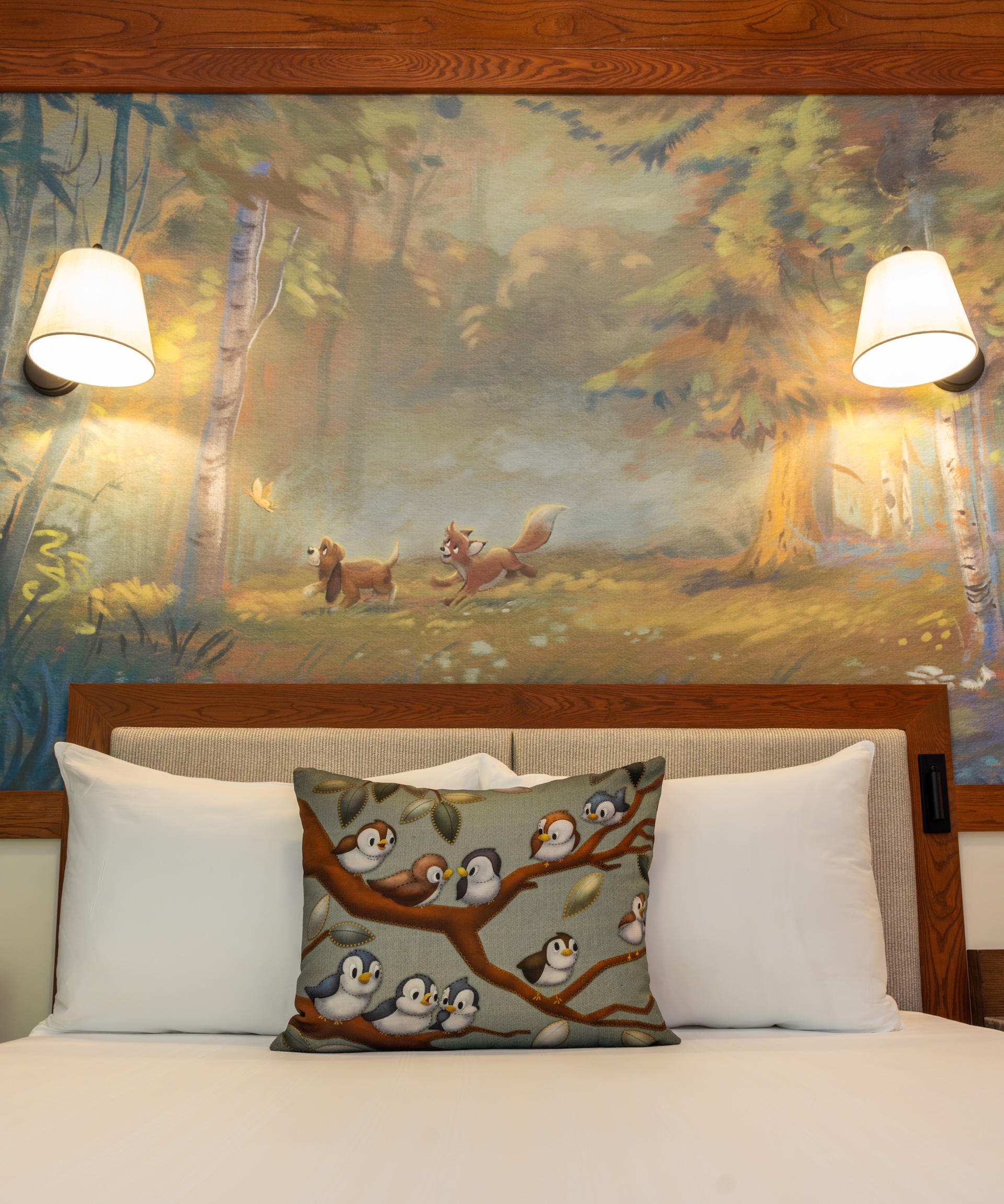 2026DVCReimaginedTreehouseVillas1 | The Main Street Mouse Bed with White Pillows and a Decorative Penguin print Cushion on a Branch Set Against a Forest Mural and Warm Wall Lamps