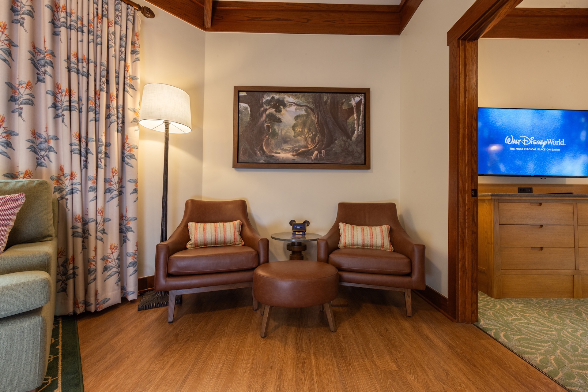 2026DVCReimaginedTreehouseVillas17 | The Main Street Mouse Two Brown Leather Chairs with a Round Ottoman Between Them in a Cozy Lounge with a Side Table and Lamp and a Forest Painting on the Wall