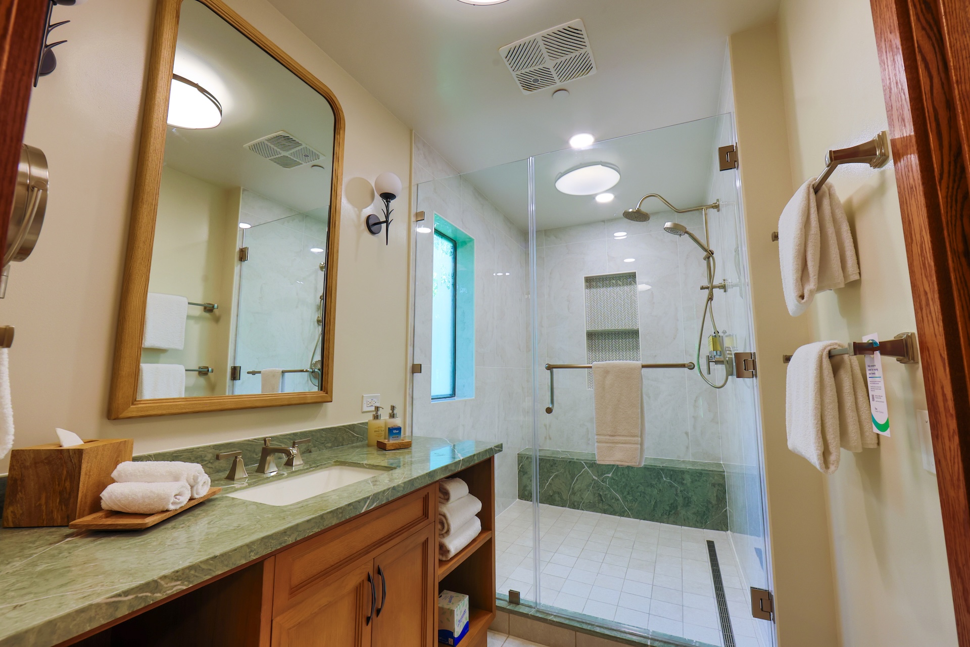 2026DVCReimaginedTreehouseVillas8 | The Main Street Mouse Bathroom with Green Marble Countertop Wooden Vanity and a Large Framed Mirror Glass Shower Enclosure with Built in Shelves and Multiple Towels on Racks